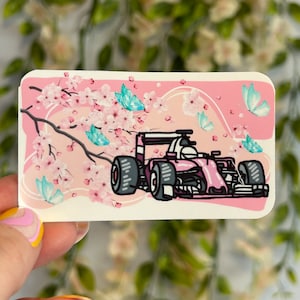 Puede incluir: Pegatina rosa y blanca con un coche de carreras rosa conduciendo por un campo de cerezos en flor. El coche tiene un casco de piloto blanco y mariposas azules vuelan alrededor del coche.