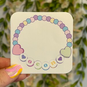 Puede incluir: Una pegatina blanca con un collar de cuentas de colores pastel con un corazón en cada extremo. Las cuentas deletrean la palabra "Bienvenido" en letras de colores.