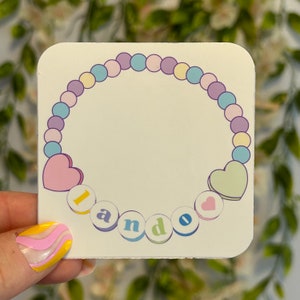 Peut inclure: Un autocollant carré avec un motif de bracelet de perles aux couleurs pastel. Le bracelet entoure un espace vide et comporte deux breloques en forme de cœur. Le mot "I and O" est épelé en lettres colorées en dessous.