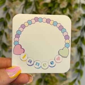 Op de afbeelding: Een witte sticker met een pastelkleurige kralenketting met harten en het woord "lance" gespeld in kleurrijke letters.