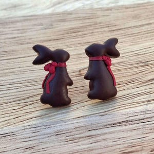 May include: A pair of dark brown bunny-shaped earrings with red ribbon detailing. The earrings are set against a light brown wooden surface. The bunny earrings have a bow around their necks, adding a festive touch.