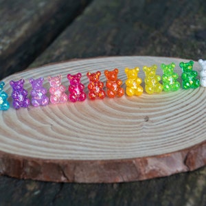 Op de afbeelding: Een regenboogassortiment van oorbellen in de vorm van gummyberen. De oorbellen zijn in een rij op een houten oppervlak gerangschikt. De kleuren van de oorbellen zijn blauw, paars, roze, rood, oranje, geel, groen en wit.