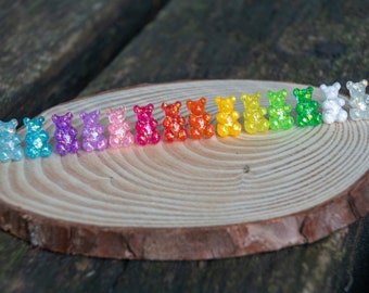 May include: A rainbow assortment of gummy bear shaped earrings. The earrings are arranged in a line on a wooden surface. The colors of the earrings are blue, purple, pink, red, orange, yellow, green, and white.