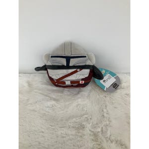 May include: A plush toy shaped like the Mandalorian's helmet and armor. The helmet is light gray with a dark blue visor, and the armor is a mix of gray, brown, and maroon. A Star Wars tag is attached.