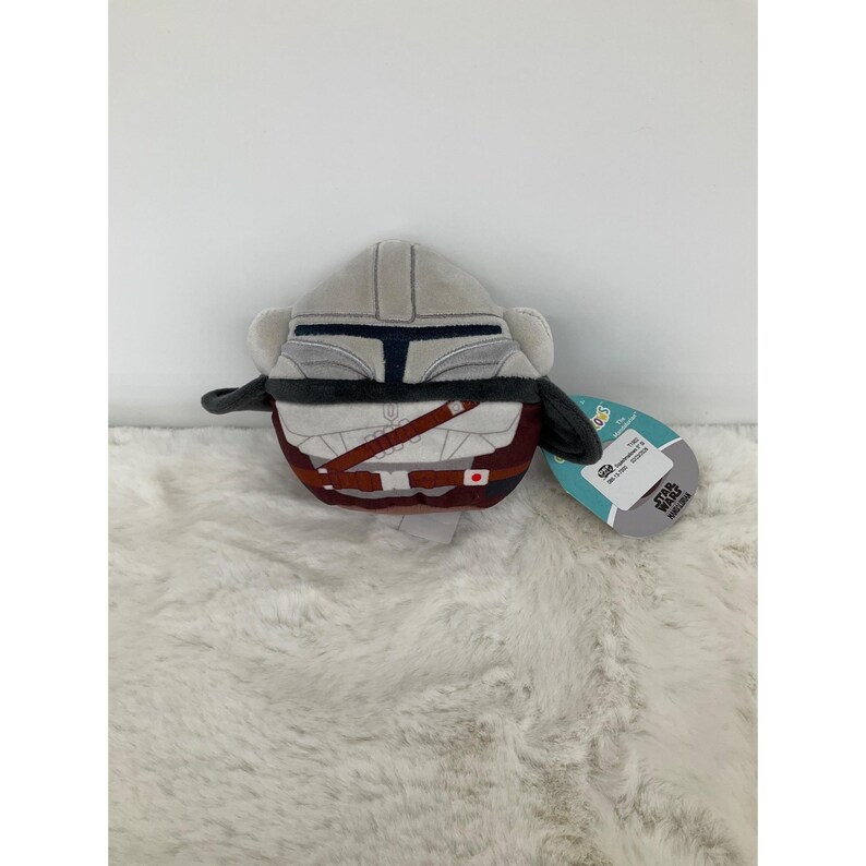May include: A plush toy shaped like the Mandalorian's helmet and armor. The helmet is light gray with a dark blue visor, and the armor is a mix of gray, brown, and maroon. A Star Wars tag is attached.