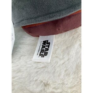 May include: Close-up of a plush toy with a gray top, burgundy middle section, and a white tag. The tag features the "STAR WARS" logo in black text. The toy is resting on a white, fluffy surface.