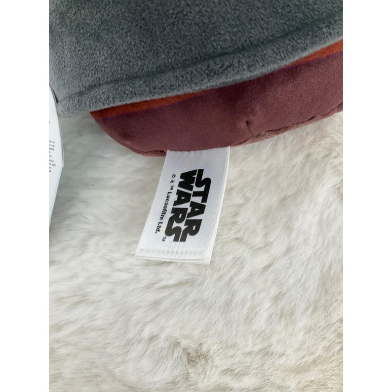 May include: Close-up of a plush toy with a gray top, burgundy middle section, and a white tag. The tag features the "STAR WARS" logo in black text. The toy is resting on a white, fluffy surface.