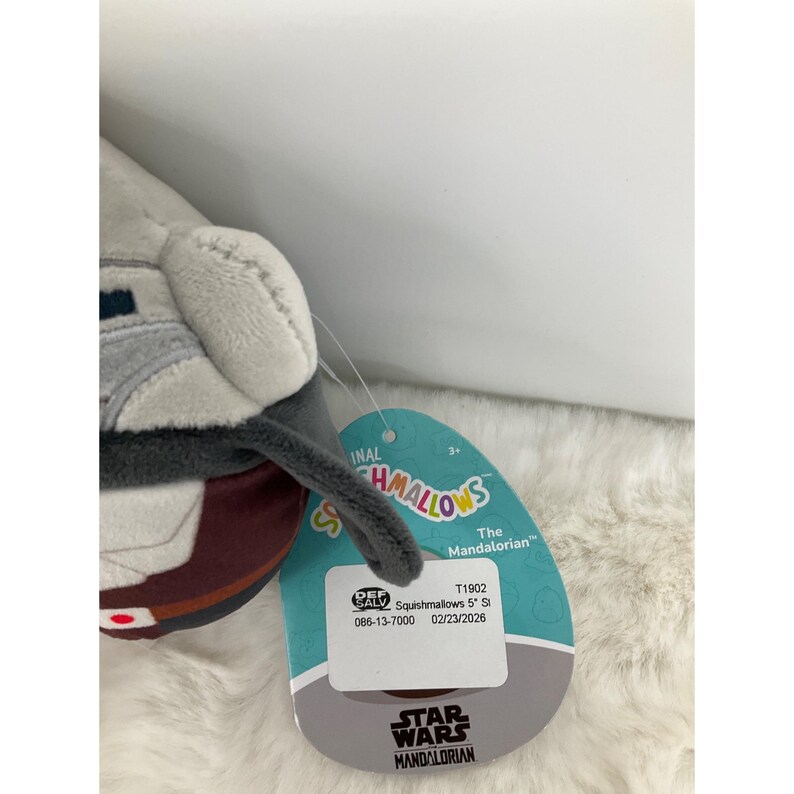 May include: A plush toy of The Mandalorian character from Star Wars. The toy is primarily gray, brown, and white, with a tag that reads "Squishmallows" and "Star Wars Mandalorian."