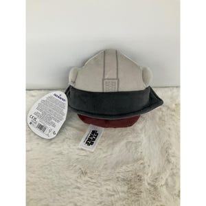 May include: A plush toy helmet in shades of gray, dark gray, and maroon. The helmet has a detailed design with a dark gray visor and ear pieces. A Star Wars tag is attached.