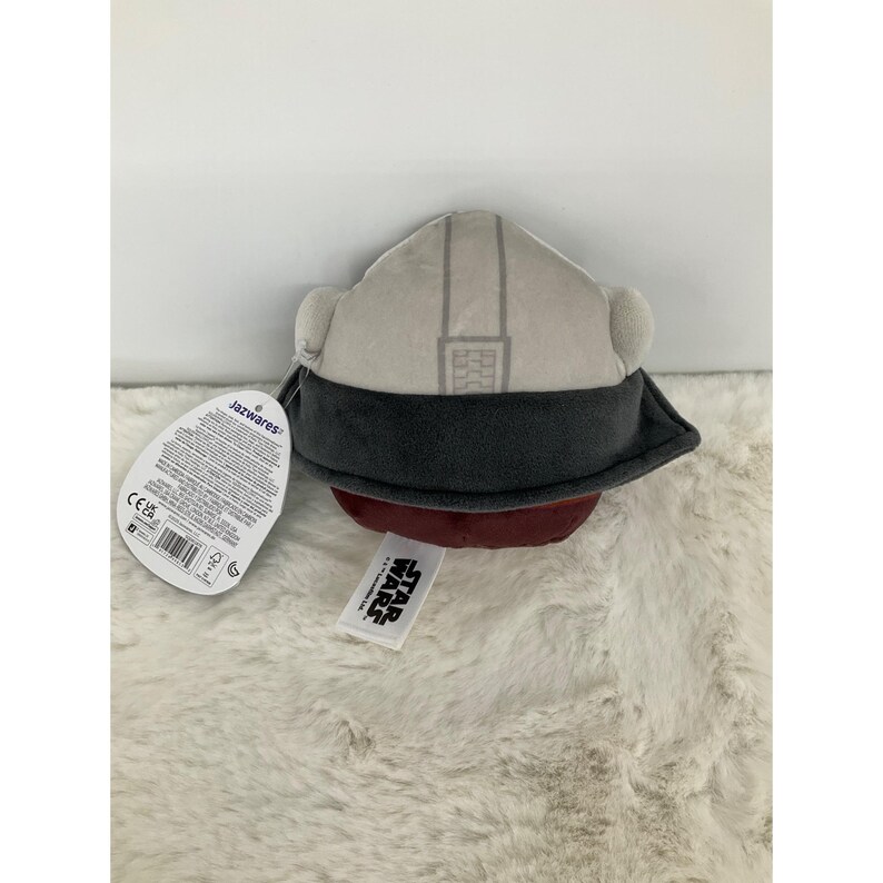 May include: A plush toy helmet in shades of gray, dark gray, and maroon. The helmet has a detailed design with a dark gray visor and ear pieces. A Star Wars tag is attached.