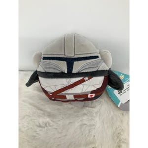 May include: A plush toy shaped like a Mandalorian helmet and armor. The toy is primarily gray with black and brown accents, and a white torso. It has a tag attached with text.