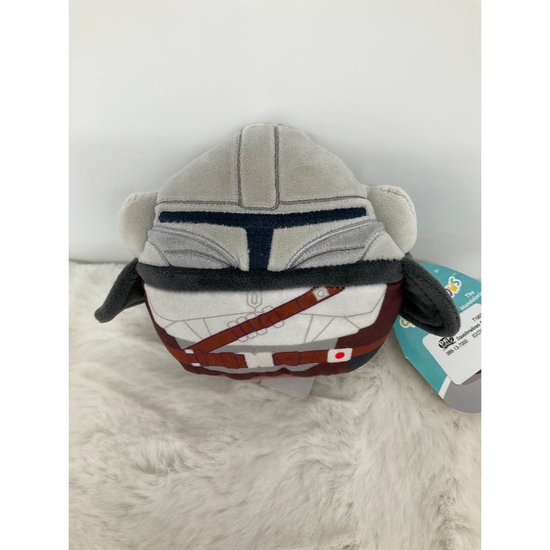 May include: A plush toy shaped like a Mandalorian helmet and armor. The toy is primarily gray with black and brown accents, and a white torso. It has a tag attached with text.