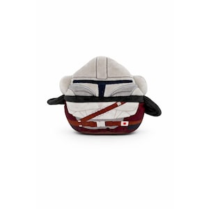 May include: A plush toy shaped like a Mandalorian helmet and armor. The toy features a gray helmet, black accents, and a burgundy and white body with brown straps. The toy has small black appendages on either side.
