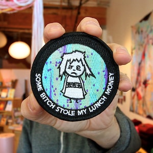 May include: A black and white embroidered patch with a cartoon character and the text "Some Bitch Stole My Lunch Money".
