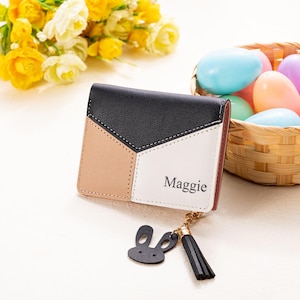 May include: A tri-color wallet with black, tan, and white geometric panels. The wallet has the name "Maggie" printed on it and a small black bunny charm with a tassel. A basket of Easter eggs and yellow flowers are in the background.