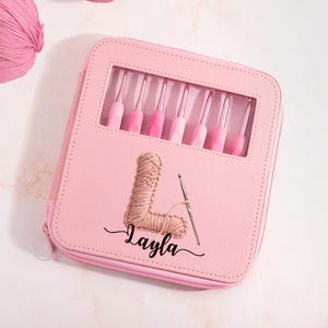 May include: A light pink square zippered case featuring a clear rectangular window displaying seven crochet hooks with pink handles. The front is printed with a large letter L made of yarn, a silver crochet hook, and the name Layla in black script.