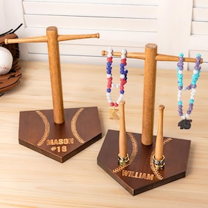 Puede incluir: Dos expositores de joyas de madera con temática de béisbol. Cada expositor tiene una base de home plate con costuras de béisbol grabadas y texto personalizado. Un expositor contiene un collar y un pequeño portaanillos con forma de bate. El otro contiene un collar y un anillo.