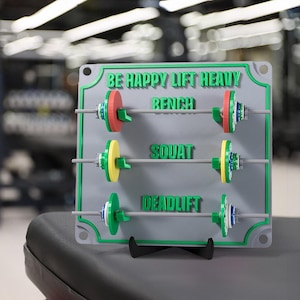 Peut inclure: Une pancarte de motivation grise et verte pour une salle de sport ou un espace d'entraînement à domicile. Elle présente une barre d'haltères avec des plaques de poids qui peuvent être déplacées pour suivre les progrès des exercices de développé couché, de squat et de soulevé de terre. La pancarte indique "Be Happy Lift Heavy" et comprend les noms des exercices : "Bench", "Squat" et "Deadlift".