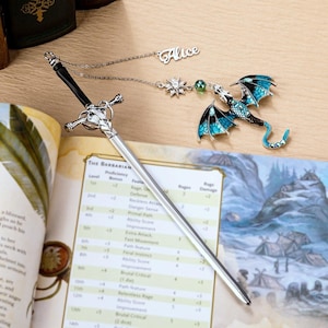 Può includere: Un segnalibro a forma di spada argentata con un'impugnatura nera e un ciondolo a forma di drago con ali blu. È attaccata una catena con il nome "Alice". Il segnalibro è su un tavolo accanto a un libro aperto con testo.