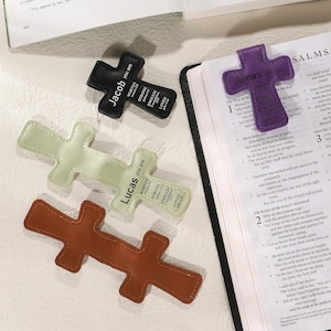May include: A collection of cross-shaped bookmarks in various colors, including black, purple, light green, and brown. Each bookmark features a name and inspirational words. One is placed on an open book.