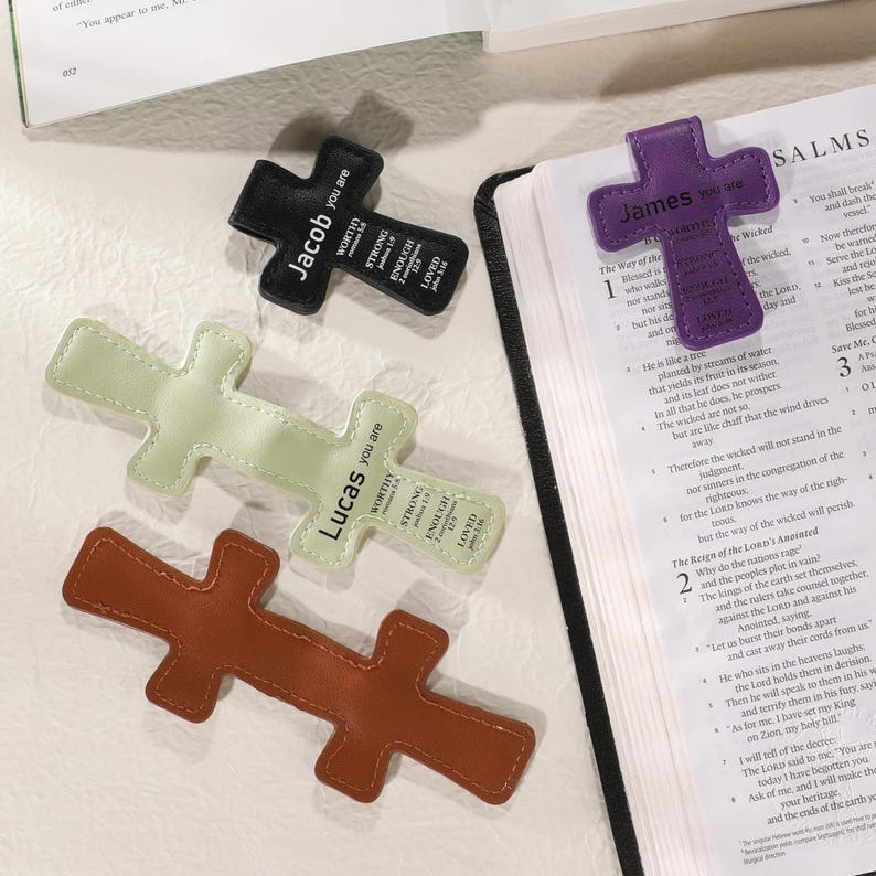 May include: A collection of cross-shaped bookmarks in various colors, including black, purple, light green, and brown. Each bookmark features a name and inspirational words. One is placed on an open book.