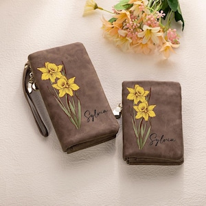 Puede incluir: Dos carteras marrones, una rectangular con correa de muñeca y otra cuadrada plegable, ambas con diseños florales de narcisos amarillos y el nombre "Sylvia". Un ramo de flores en el fondo. Las carteras están hechas de un material texturizado.