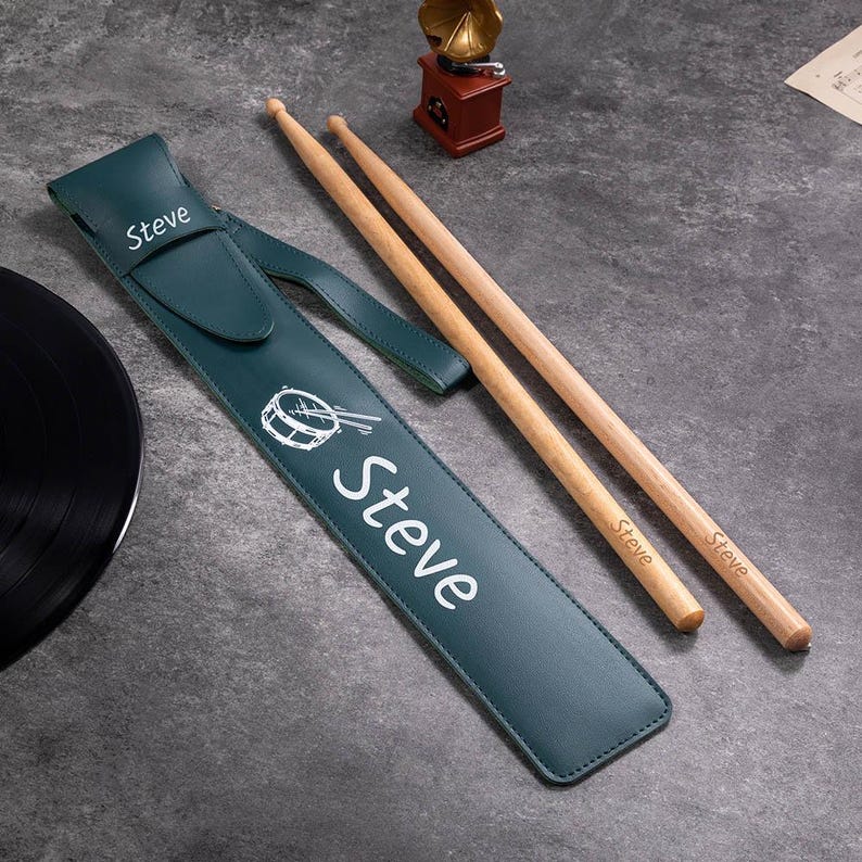 May include: A green leather drumstick case with the name "Steve" printed on it. The case has a small pocket on the top and a white illustration of a drum. Two wooden drumsticks are laying next to the case, each with the name "Steve" printed on them.