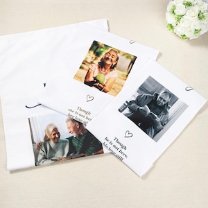 May include: White pillowcases with printed photographs of elderly people. One photo shows a woman holding a green fruit, another a black and white photo of a man reading. Text includes "Though she is not her, her love still" and "Though he is not here, his love still."
