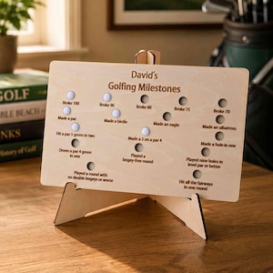 Könnte beinhalten: Eine helle Holztafel mit der Aufschrift David's Golfing Milestones und kreisförmigen Aussparungen für Golfbälle. Das Schild listet Erfolge wie Broke 100, Made a birdie und Hit all the fairways in one round auf und steht auf einem Holzständer.
