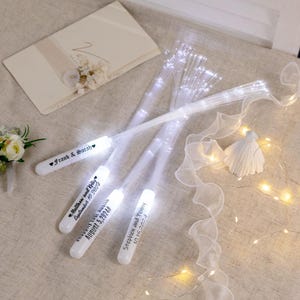 May include: Four white plastic wands with fiber optic lights and personalized messages for a wedding. The messages include the names of the couples and their wedding dates.  The wands are lying on a white surface with a white fabric and string lights.