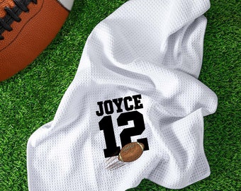 May include: White football towel with the name "JOYCE" and the number "12" in black. A football graphic is printed below the number. An American football is in the background, resting on green turf.