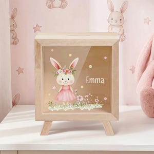 Könnte beinhalten: Holz-Schattenbox mit klarer Front, mit einer Hasen-Illustration mit Blumenkrone und rosa Kleid. Der Name "Emma" ist auf der Vorderseite aufgedruckt. Die Box steht auf einer kleinen Holzstaffelei, mit einer rosa Tapete mit Hasen und Sternen im Hintergrund.