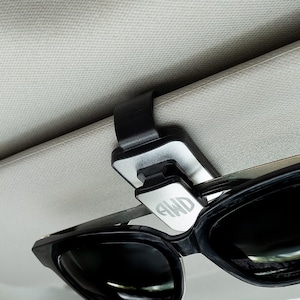 May include: A silver car visor clip with a black strap and the letters "AWD" engraved on the front. The clip is holding a pair of black sunglasses.