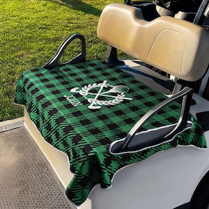 Peut inclure: Une housse de siège de voiturette de golf avec un motif à carreaux vert et noir. La housse présente un emblème blanc avec des clubs de golf, une raquette de tennis et le nom "JOHN".