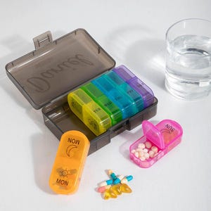 May include: A clear plastic pill organiser with seven compartments labelled for each day of the week in rainbow colours. An orange pill case is labelled "NOW" and "MON". A pink pill case is open, filled with pills. A glass of water is in the background.