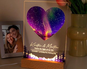 Op de afbeelding: Een gepersonaliseerde acryl nachtlamp met een hartvormig ontwerp met een sterrenhemel en noorderlicht. De lamp heeft een houten voet en bevat de namen "Kirsten & Martin" en de tekst "You're the light of my life" samen met een datum en coördinaten.