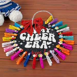 Può includere: Un cartello bianco e nero con il testo "My Cheer Era" circondato da mollette colorate con frasi motivazionali. Il cartello ha un fiocco di glitter rosso e un manico argentato.