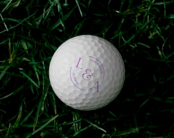 May include: A white golf ball with a dimpled surface, resting on green grass. The ball features a purple design with the initials "L & A", the date "07.15.2024", and the names "Louie & Amelia".
