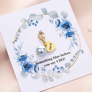 May include: A gold-colored charm with a light blue pearl and a heart-shaped tag with the initials "A&D" is attached to a card. The card features a floral wreath with blue flowers and the text "Something blue before you say I DO!"