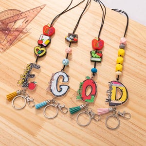 Pode incluir: Cinco cordões coloridos para crachás de identificação de professores com chaveiros. Cada cordão apresenta o nome de um professor diferente, uma letra do alfabeto e um amuleto com tema escolar. Os amuletos incluem uma maçã, um livro, um lápis e uma minhoca.