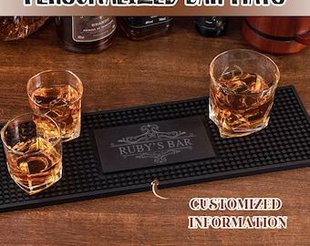May include: Black bar mat with a personalized inscription "Ruby's Bar" in a decorative font. The mat has a textured surface and is designed to protect countertops from condensation and spills. Three glasses of amber liquid are placed on the mat.
