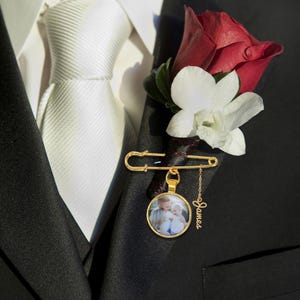 Könnte beinhalten: Goldfarbene Anstecknadel mit einem kleinen, runden Anhänger mit einem Foto von zwei Kindern. Die Nadel ist an einem schwarzen Revers befestigt, neben einer roten Rose und einer weißen Blumen-Boutonniere. Ein kleiner Anhänger mit dem Namen "James" hängt am Anhänger.