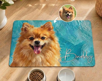 Puede incluir: Una alfombrilla rectangular para mascotas con un retrato pintado de un perro Pomerania naranja esponjoso con la lengua fuera sobre un fondo azul verdoso texturizado. El nombre Buddy está escrito en letra cursiva blanca junto a una foto circular.