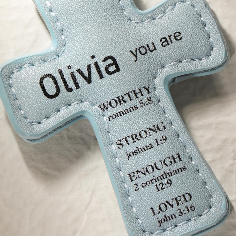 May include: A light blue cross-shaped item with the name "Olivia" and the words "you are" printed in black. The cross also features the words "WORTHY," "STRONG," "ENOUGH," and "LOVED" with associated Bible verses.