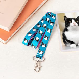 Puede incluir: Un cordón turquesa con un patrón repetido de caras de gatos en blanco y negro. El cordón tiene un cierre de metal plateado y un reverso rosa. Una fotografía de un gato blanco y negro está en el fondo.