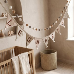 May include: A decorative bunting banner featuring triangular fabric flags in cream and beige, each displaying a brown embroidered letter to spell the name LEONARDO, accented with small white pom-poms and a matching felt ball garland string.