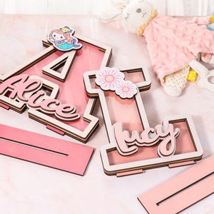May include: Two pink wooden letters, A and I, with the names "Alice" and "Lucy" written on them. The letters are layered and have a white border. The letter A has a mermaid figurine on top. The letters are on a pink surface.
