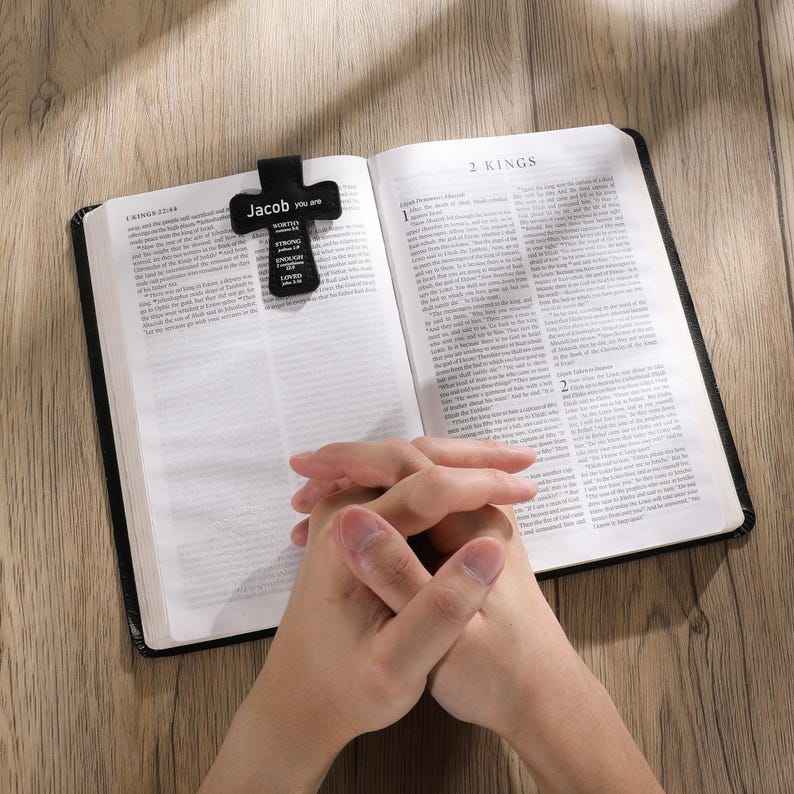 May include: An open book with the text "2 Kings" visible, featuring a black cross-shaped bookmark. The bookmark has the name "Jacob" and other words printed on it. The person's hands are clasped in prayer over the open book.