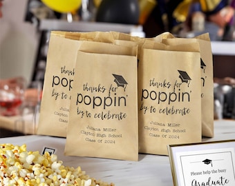 May include: Brown paper bags with black text that reads "thanks for poppin' by to celebrate" with a graduation cap icon. The bags are filled with popcorn and are used as party favors for a graduation party.