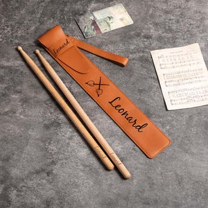May include: A brown leather drumstick case with the name "Leonard" engraved on it. The case is open and two wooden drumsticks are inside. The drumsticks are also engraved with the name "Leonard".